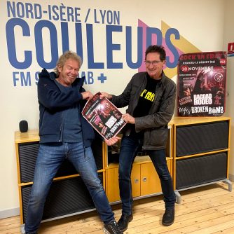 Dans les studios de Radio Couleurs, devant le logo inscrit sur le mur, deux hommes font semblant de se disputer l'affiche de Rock'in Frat. L'homme sur la droite, aux cheveux gris, porte un haut bleu foncé, une polaire bleu marine, un jean et des baskets noires et blanches. L'homme sur la gauche, aux cheveux noirs, porte des lunettes, un t-shirt noir sur lequel on devine, en jaune, l'inscription "Rock'in Frat", un blouson en cuir marrob foncé, un jean et des chaussures en cuir. Les deux hommes sourient à pleines dents.