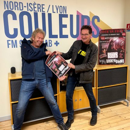 Dans les studios de Radio Couleurs, devant le logo inscrit sur le mur, deux hommes font semblant de se disputer l'affiche de Rock'in Frat. L'homme sur la droite, aux cheveux gris, porte un haut bleu foncé, une polaire bleu marine, un jean et des baskets noires et blanches. L'homme sur la gauche, aux cheveux noirs, porte des lunettes, un t-shirt noir sur lequel on devine, en jaune, l'inscription "Rock'in Frat", un blouson en cuir marrob foncé, un jean et des chaussures en cuir. Les deux hommes sourient à pleines dents.