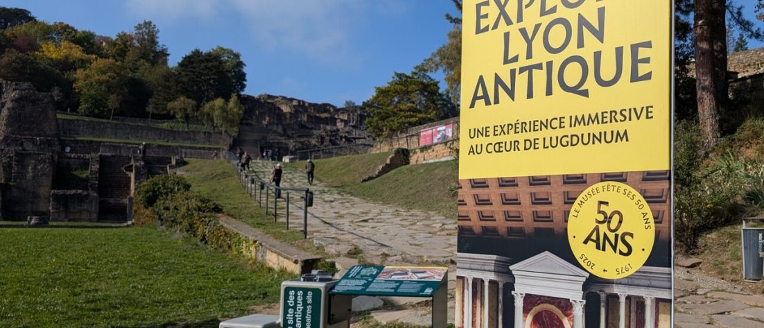 Photo de l'entrée de l'expérience Explore Lyon Antique. Au fond, on y voit beaucoup de verdure. Des arbres et une colline se détache en arrière plan. Un chemin en pierre, qui part d'en bas, grimpe le long de la colline, parré de barrières. Au début du chemin, un panneau pédagogique vert ; à côté d'un grand panneau vertical sur lequel, au dessus de la photo d'un bâtiment antique, un bandeau jaune pâle indique "Lugdunum: musée théâtres romains - EXPLORE LYON ANTIQUE - une expérience immersive au coeur de Lugdunum." Un badge de la même couleur indique également "50 ans".
