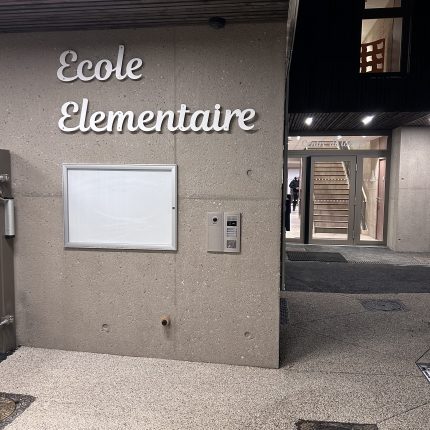 Photographie de l'entrée de l'école élémentaire de Montferrat. A gauche, un mur gris, sur lequel figurent un panneau d'affichage et un interphone. On devine aussi un portillon. Au dessus du panneau, une enseigne en jolies boucles indique "Ecole élémentaire". Sur la droite, une sorte de couloir extérieur se découpe, menant jusqu'à la porte d'entrée, derrière laquelle on distingue un grand escalier.