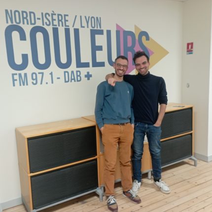 Mathias et Antoine, du groupe Lingus, posant dans le studio, devant le logo de Radio Couleurs. Antoine a son bras passé autour du cou de Mathias, il semble pointer l'objectif de l'indexe. Mathias a les deux mains dans les poches. Les deux sourient à pleines dents.