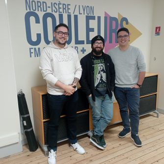Photo du groupe de métal Last Addiction devant le logo de Couleurs. Gaël, à gauche, porte un hoodie vans blanc, un jean foncé retroussé et des baskets blanches. Il est debout, les jambes légèrement écartées, sa main gauche posée sur son poignet droit. William, au centre, porte un bonnet noir, une veste assortie, un t-shirt Green Lung avec le visuel de l'album Let the Devil In (les couleurs dominantes sont le noir, le vert, et le blanc), un jean clair et des baskets noires aux semelles blanches. Il a les mains dans les poches de son jean et les jambes croisées. Dylan porte un pull gris clair aux manches retroussées, un jean foncé (mais moins que celui de Gaël), et des baskets noires. Il a des écarteurs aux oreilles. Il a également les mains dans les poches de son jean. Les trois hommes, qui portent des lunettes, regardent l'objectif en souriant.