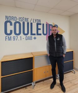 JP Barret pose dans le studio de Radio Couleurs. C'est un homme basique, aux cheveux courts gris, barbichette, lunettes. Il porte une parka noire par dessus un pull gris, un pantalon lui-même noir et des chaussures en daim asorties. 
Il pose devant un meuble, posé devant le logo de Couleurs.