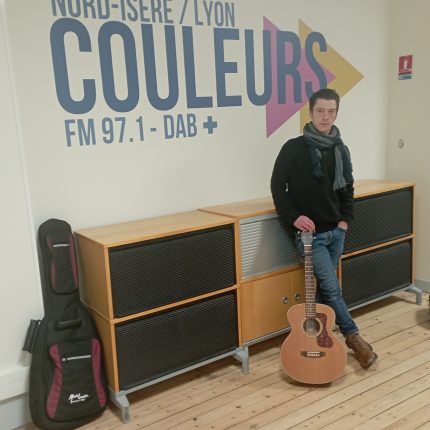 Lomnouvo, calé contre le meuble posé contre le mur sur lequel se trouve le logo de Radio Couleurs. Il a les cheveux courts, et pas de pilosité faciale apparente. Il (Lomnouvo), porte un pull noir, un jean bleu, de grosses chaussures marrons montantes ; et une écharpe grise autour du cou. Sa main droite est posée sur le haut du manche de sa guitare, qui cache son pied gauche, et sa main gauche est dans sa poche. A l'extrémité du meuble, la housse de sa guitare, bien droite.
