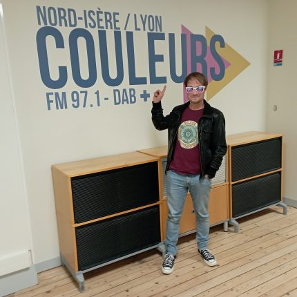 Thibault, du groupe Blue Sun Flower, devant le logo de Couleurs. Il est un poil "goofy", penché en arrière, il semble pointer le logo, un rire discret figé sur son visage, sa main gauche dans la poche de son jean. Il porte des lunettes de soleil blanches, aux verres colorés (ils semblent rose et violet foncé sur l'image). Il porte un t-shirt bordeau et un blouson de cuir.